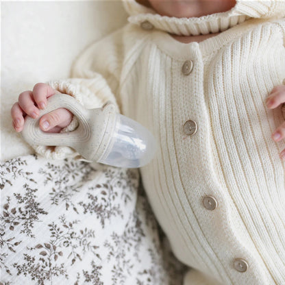 Baby in a cream knitted outfit holding a white pacifier against a soft, neutral background.